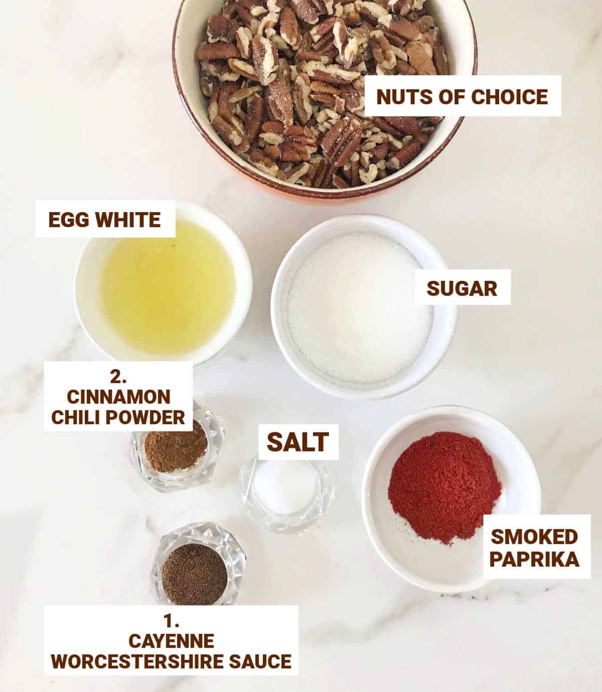 Cocktail nuts ingredients in bowls on white surface including ground spices, egg white, sugar, and salt.