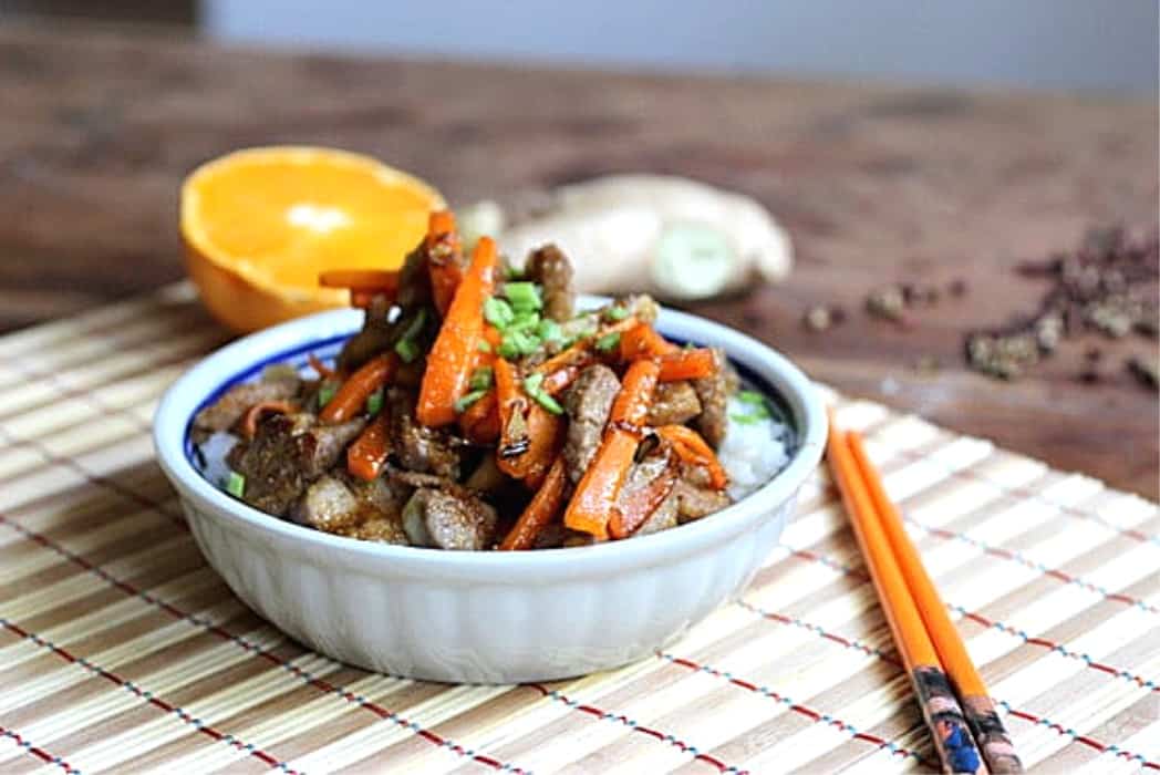 White bowl with pork and carrots, bambu placemat, orange half and chopsticks.