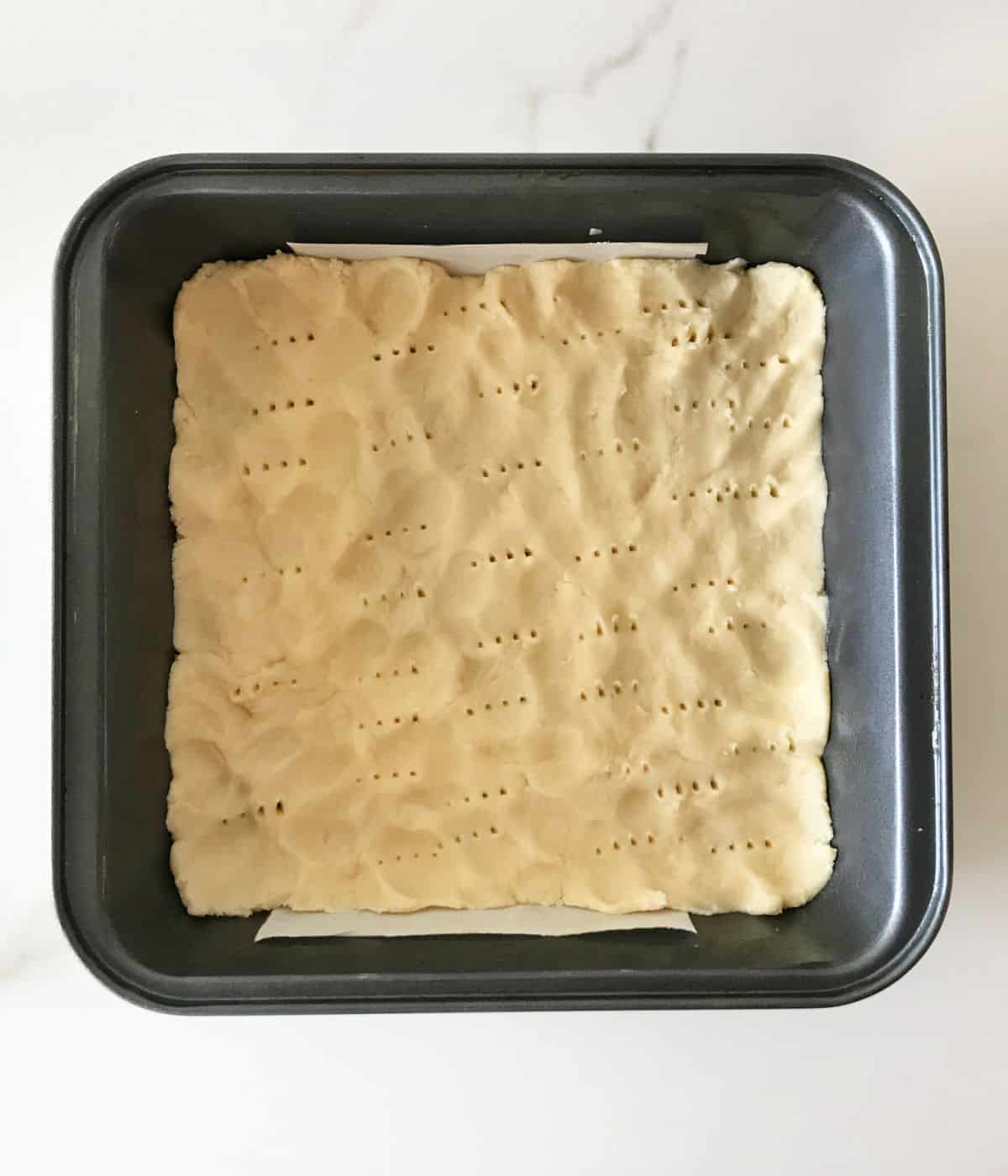 Unbaked shortbread crust pricked in a square metal baking pan on a white marble surface.