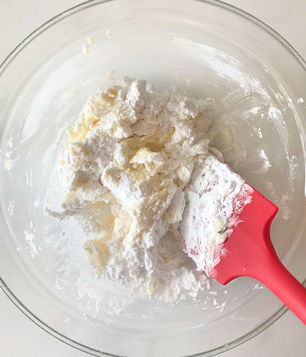 Incorporating flour into the butter mixture with a red spatula in a glass bowl. White surface.