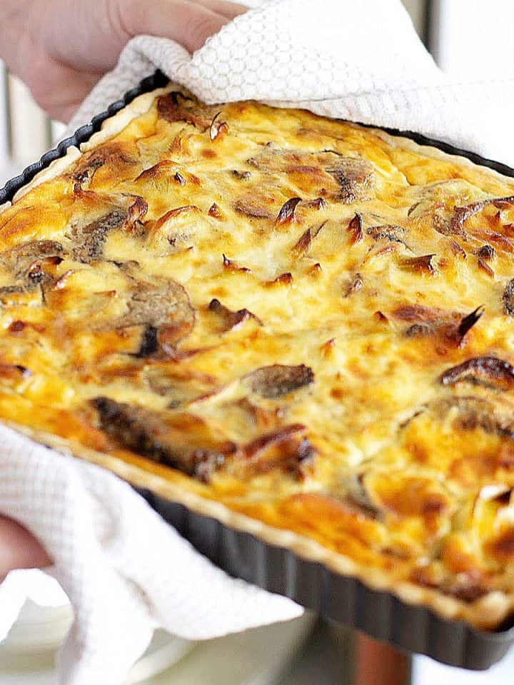 Whole baked square mushroom quiche in the pan being held with white kitchen towels.