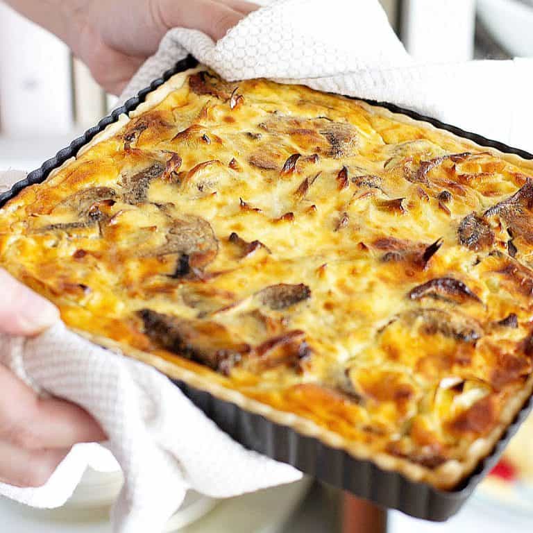 Whole baked square mushroom quiche in the pan being held with white kitchen towels.