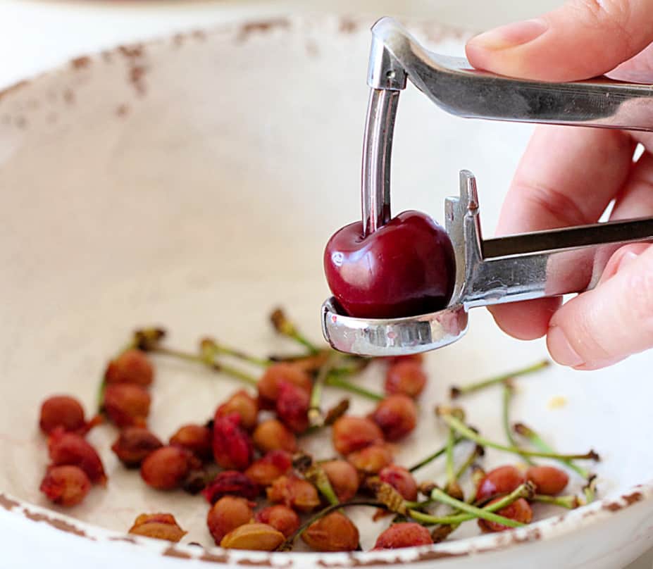 Hand with cherry pitter, whole cherry, white bowl with pits