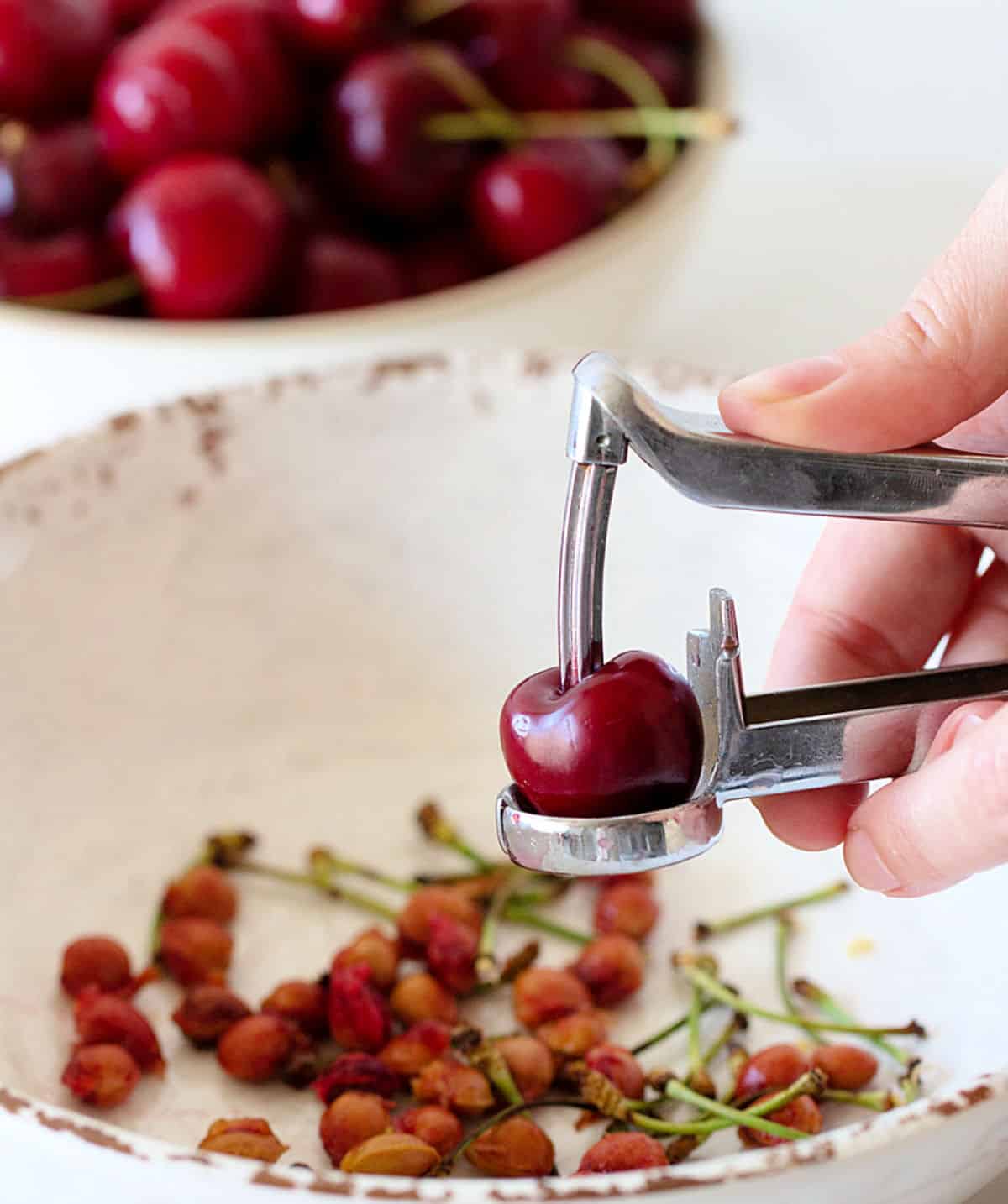 Hand pitting cherries with a cherry pitter over a white bowl.