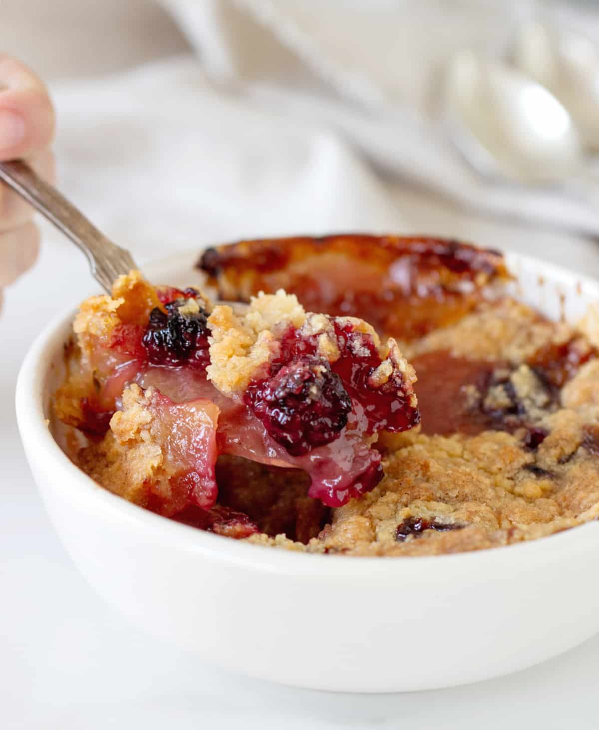 Spoon lifting apple blackberry crumble from white bowl.