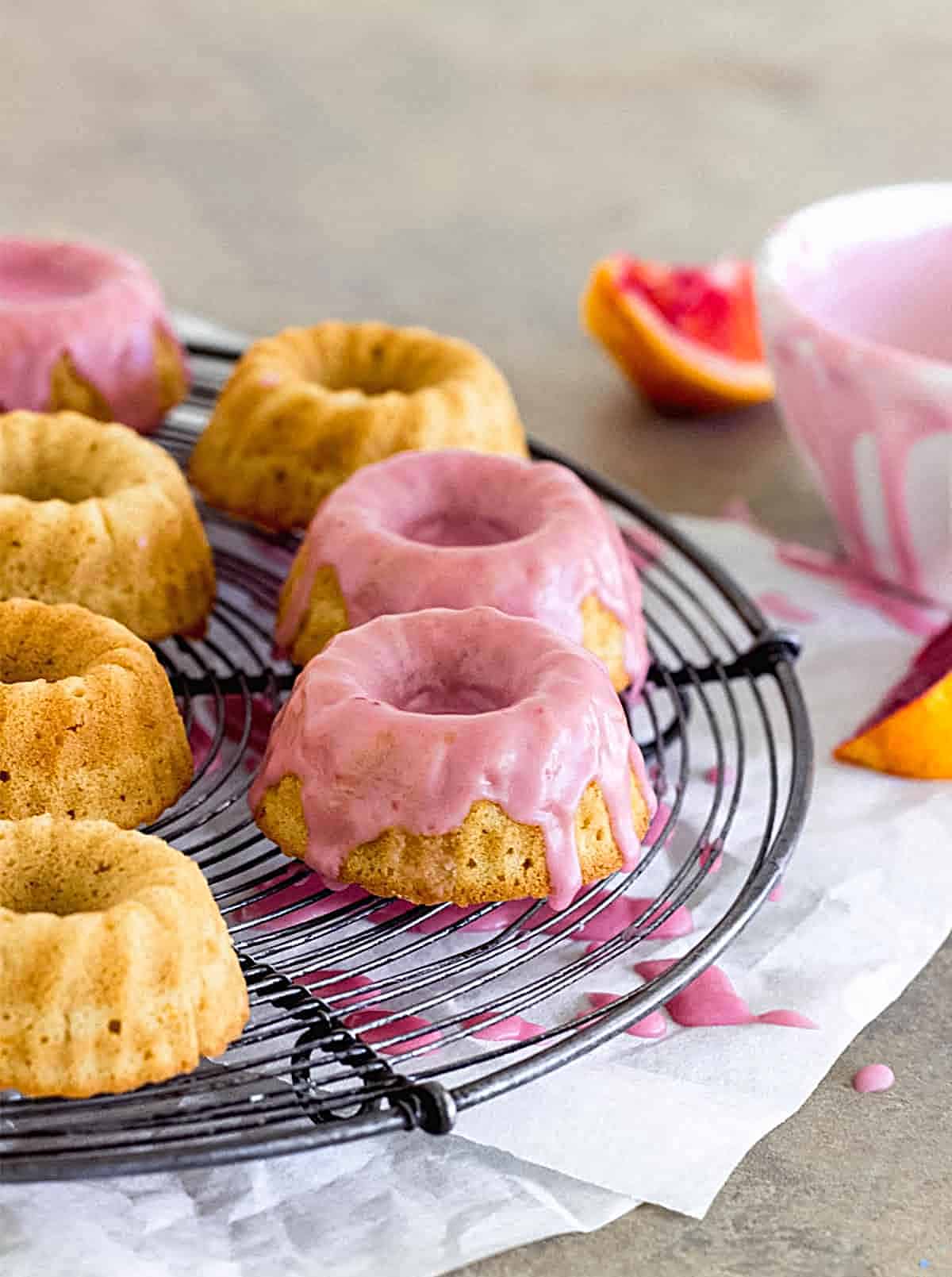 Pink Glazed mini bundt cakes on wire rack, grey surface, bowl and orange wedges on the side