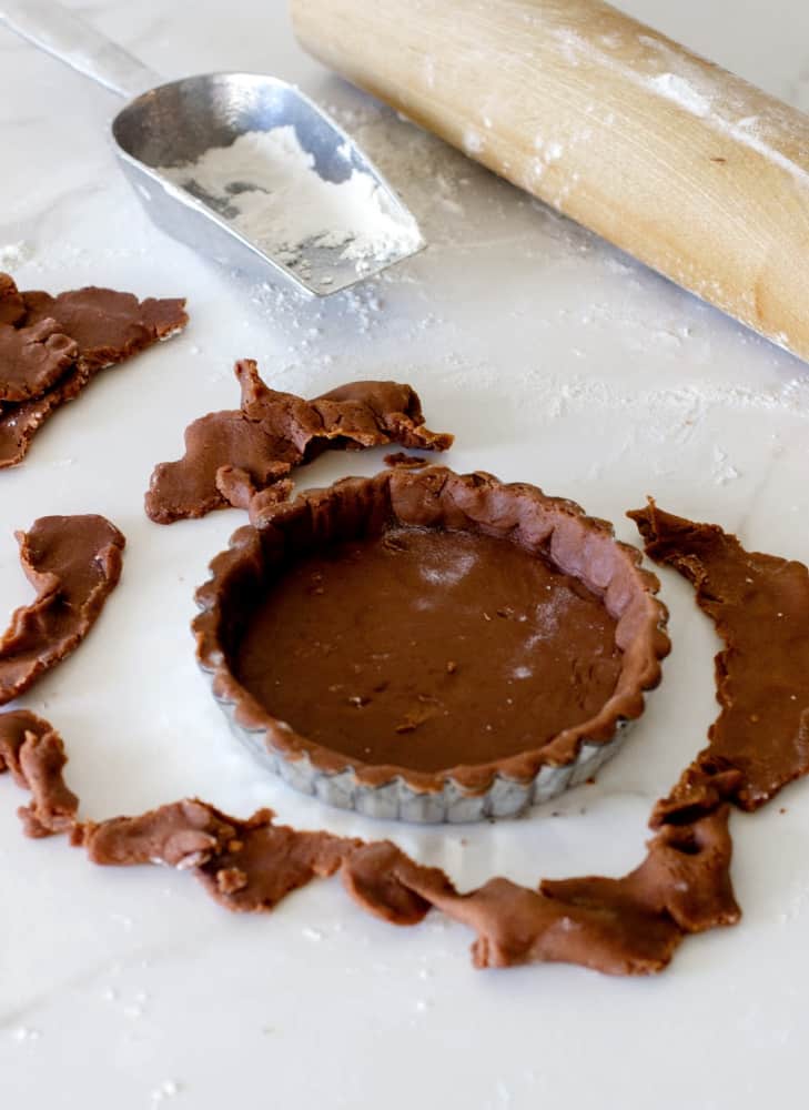 Lined individual tart pan with chocolate crust, excess pie dough, rolling pin, white surface