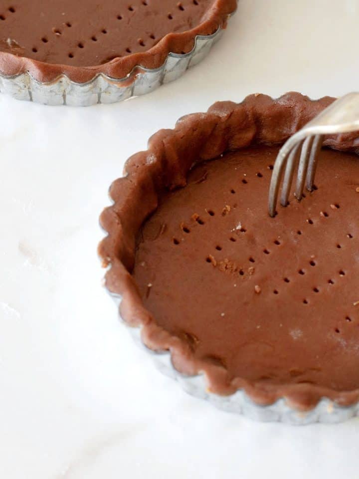 Pricking chocolate tart dough in pie tins with fork on white marble surface.