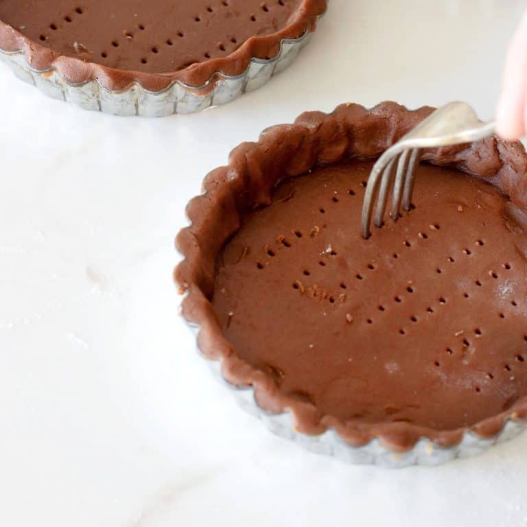 Pricking chocolate tart dough in pie tins with fork on white marble surface.