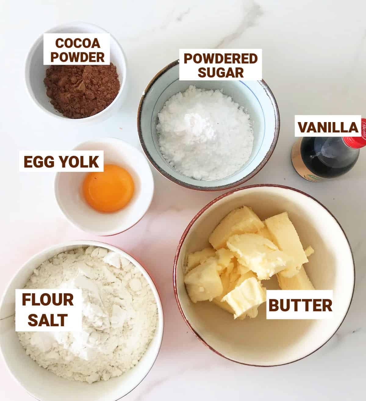 Chocolate pie crust ingredients in bowls on white surface including egg yolk, cocoa powder, vanilla, powdered sugar, butter.