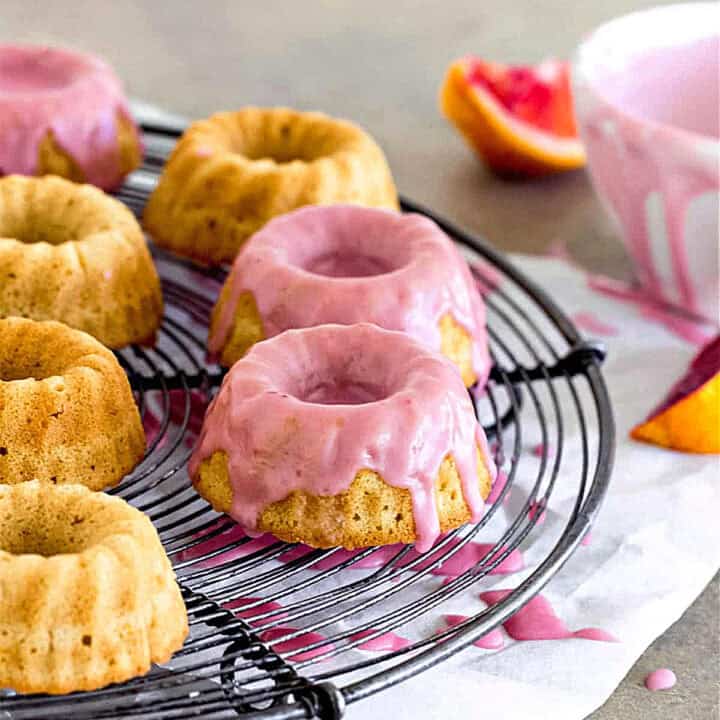 Pink glazed mini bundt cakes on a wire rack. White bowl, gray surface, white paper.