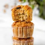 Bitten morning glory muffin on top of stack of muffins in paper liners. White background with greenery.