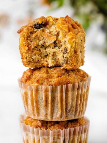 Bitten morning glory muffin on top of stack of muffins in paper liners. White background with greenery.