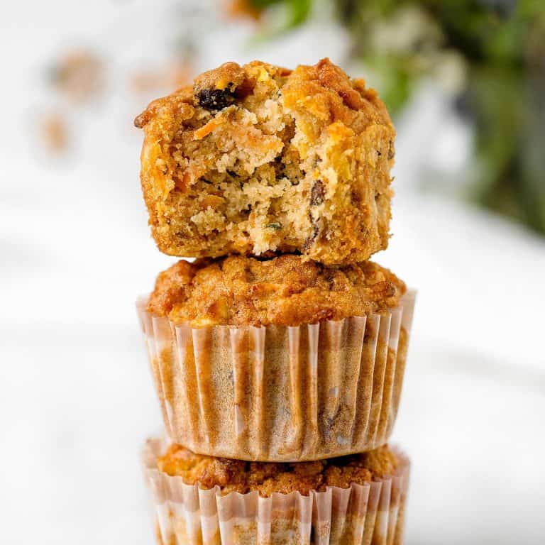 Bitten morning glory muffin on top of stack of muffins in paper liners. White background with greenery.