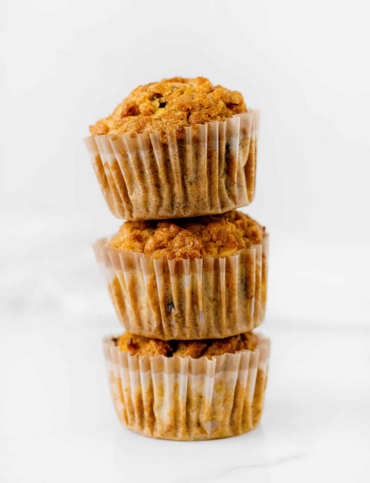 Stack of three carrot muffins in paper liner on white marble surface and background. 