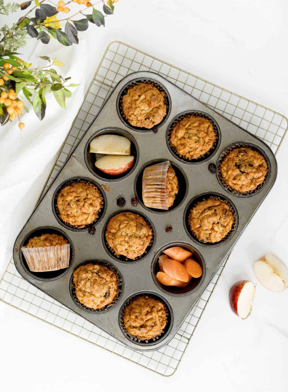 Top view of muffin pan on a wire rack with baked muffins, carrot and apple slices. White marble surface. 