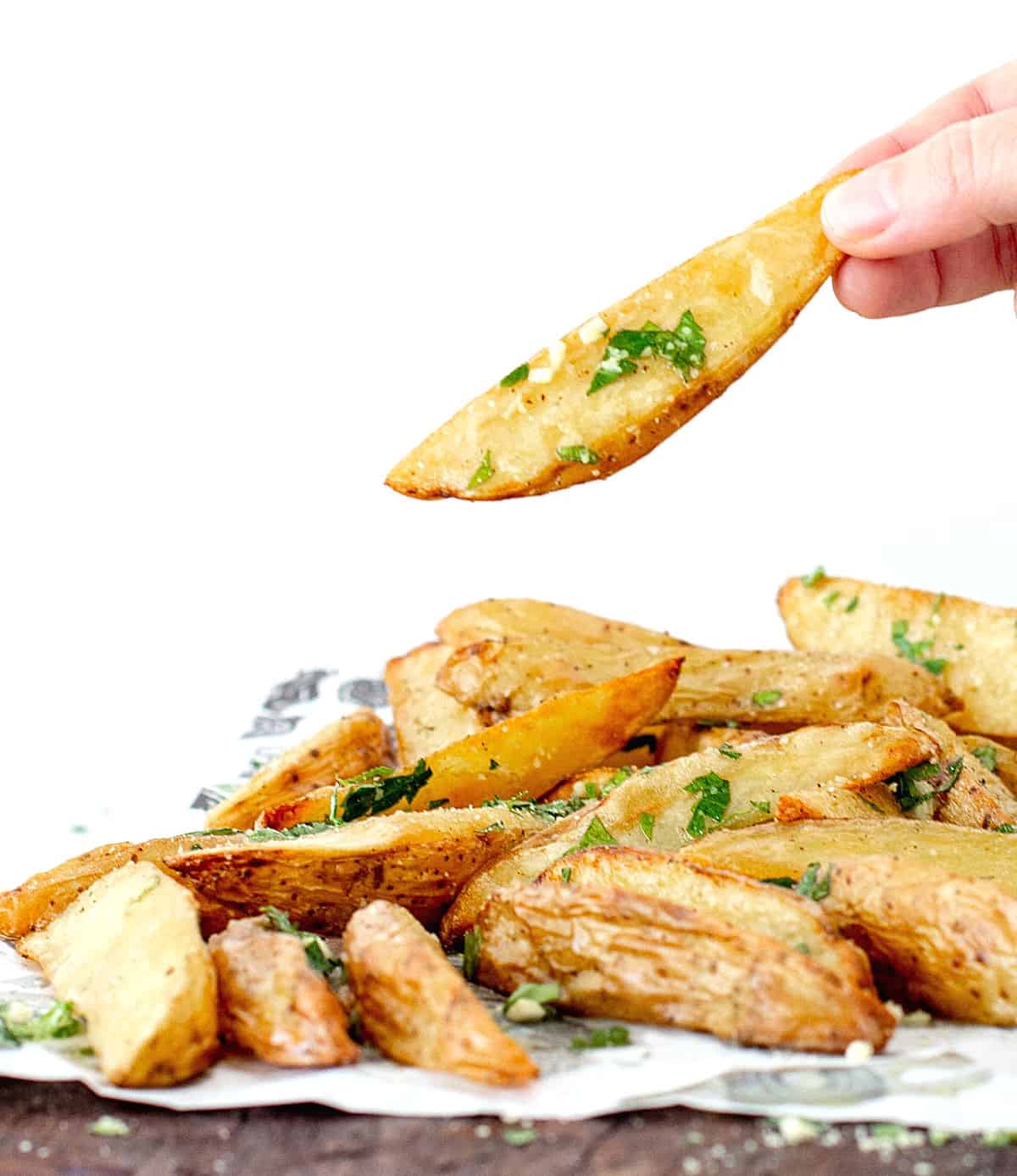 Hand holding potato wedge beneath a mound of potato wedges on a wooden board.