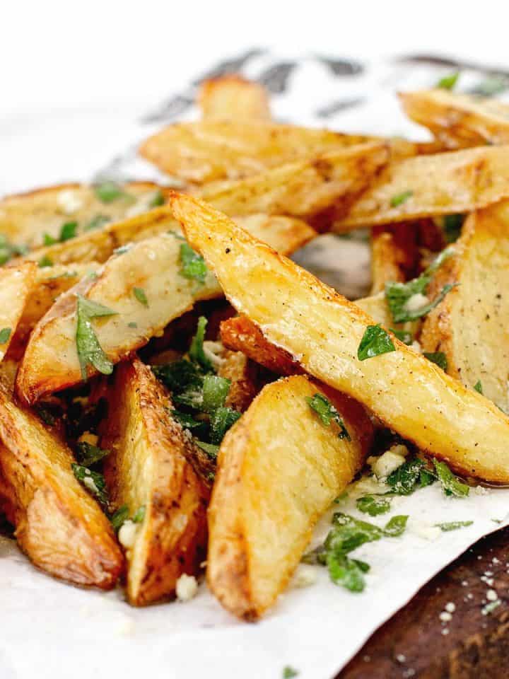 Pile of parsley and parmesan sprinkled baked potato wedges on a white piece of paper.