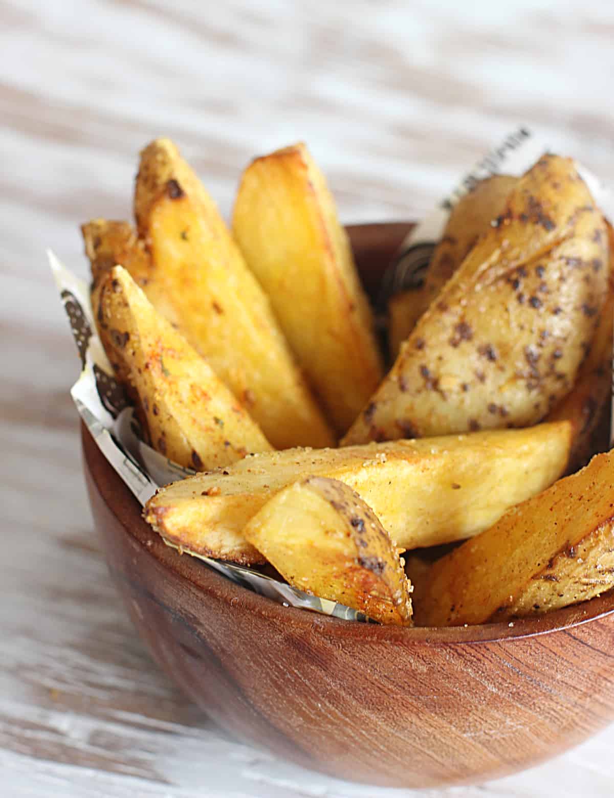 Close up image of golden potato wedges in a wooden bowl on a white-beige surface