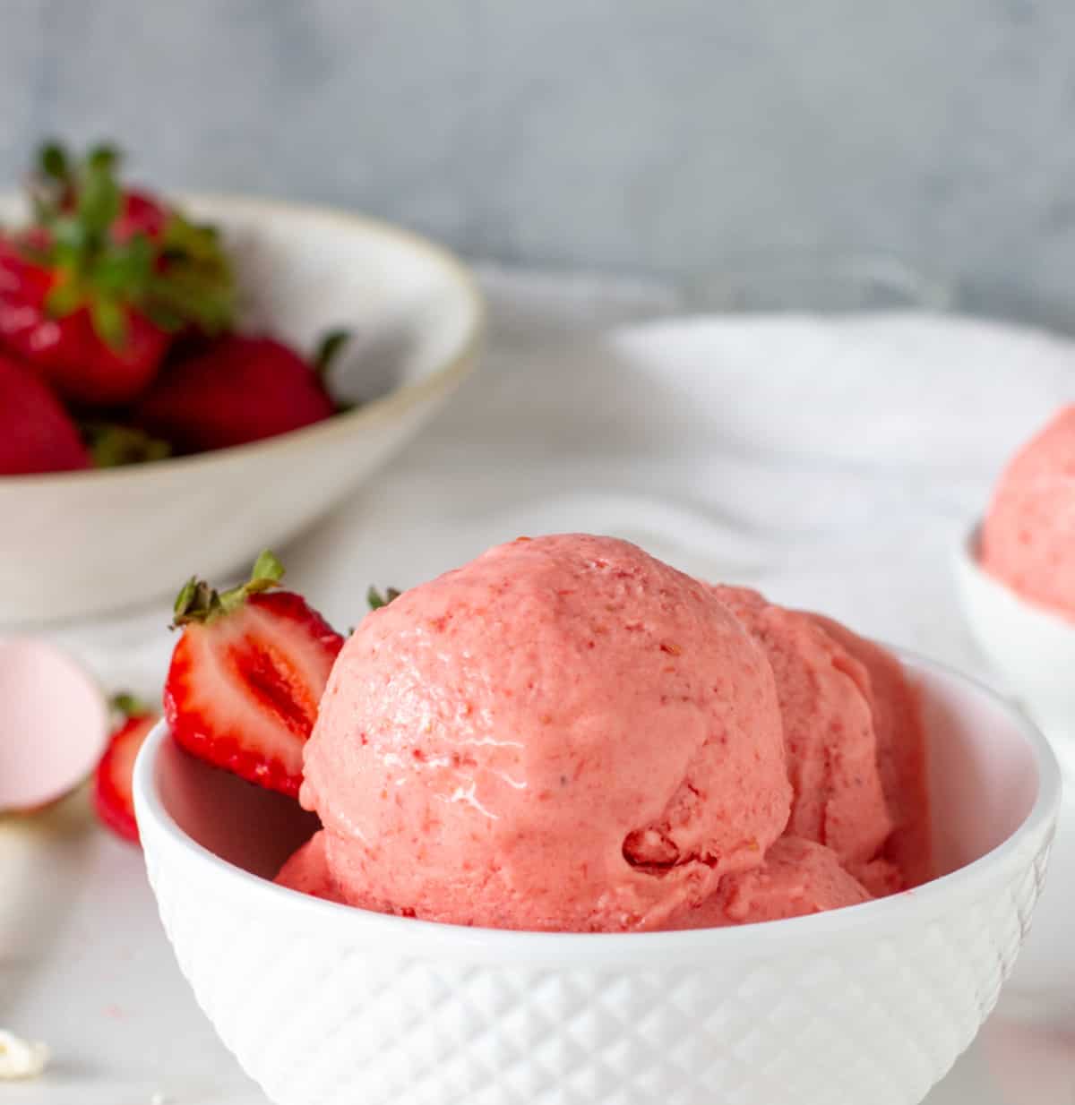 Scoops of strawberry ice cream in white bowl, strawberries beneath, a grey background