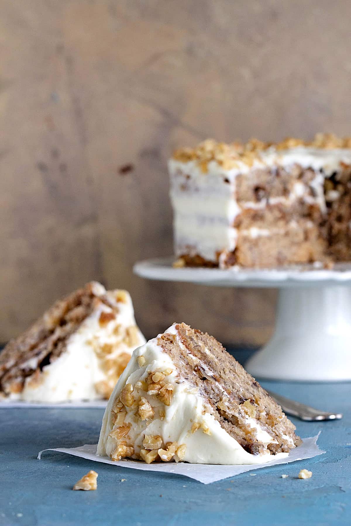 Slices of frosted layer cake with walnuts on a blue surface and brownish background with whole cake on a white cake stand.