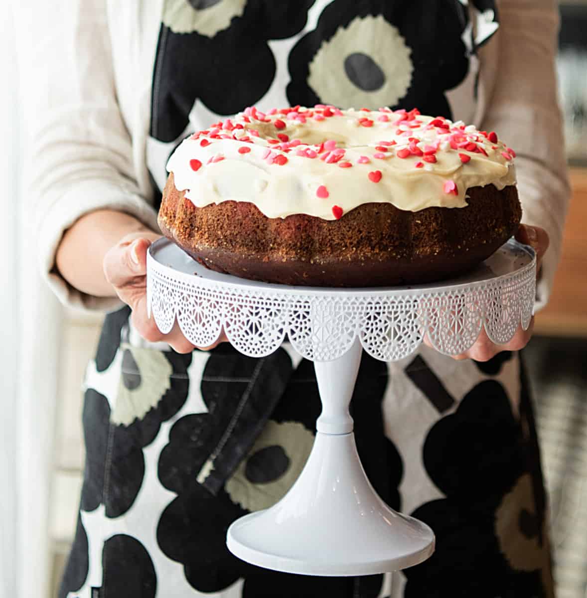 Person with black and beige apron holding white cake stand with frosted red velvet bundt