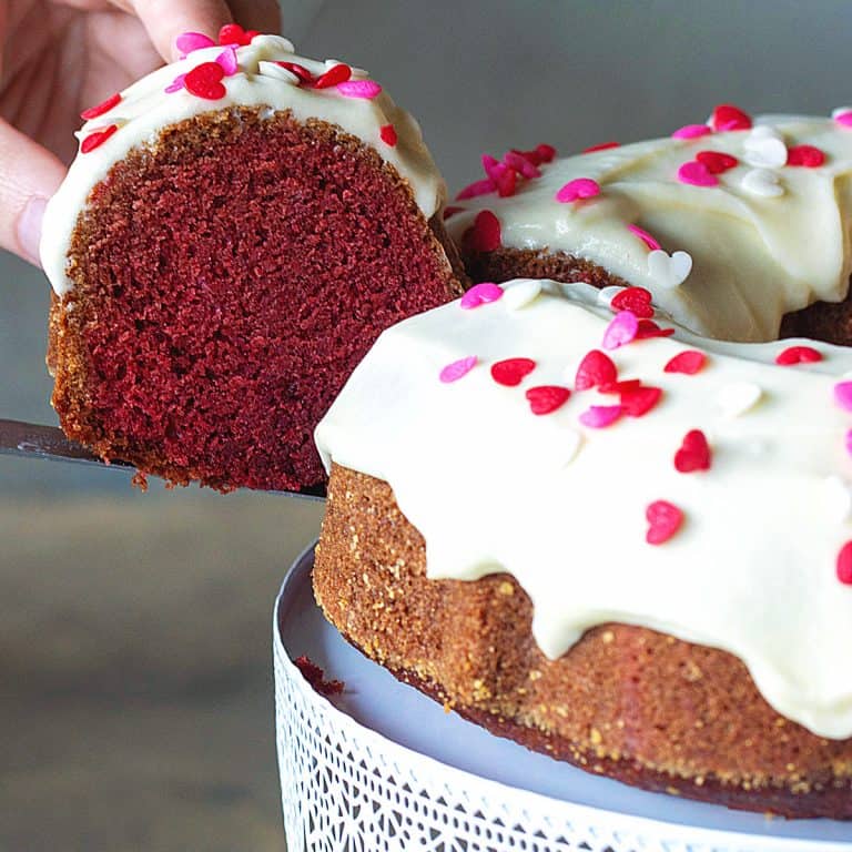 A hand lifting slice of frosted red velvet bundt cake from a white cake stand