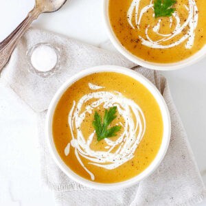 Pumpkin soup with cream drizzle on white bowls. Beige cloth on white surface.