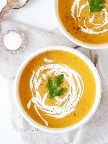 Pumpkin soup with cream drizzle on white bowls. Beige cloth on white surface.