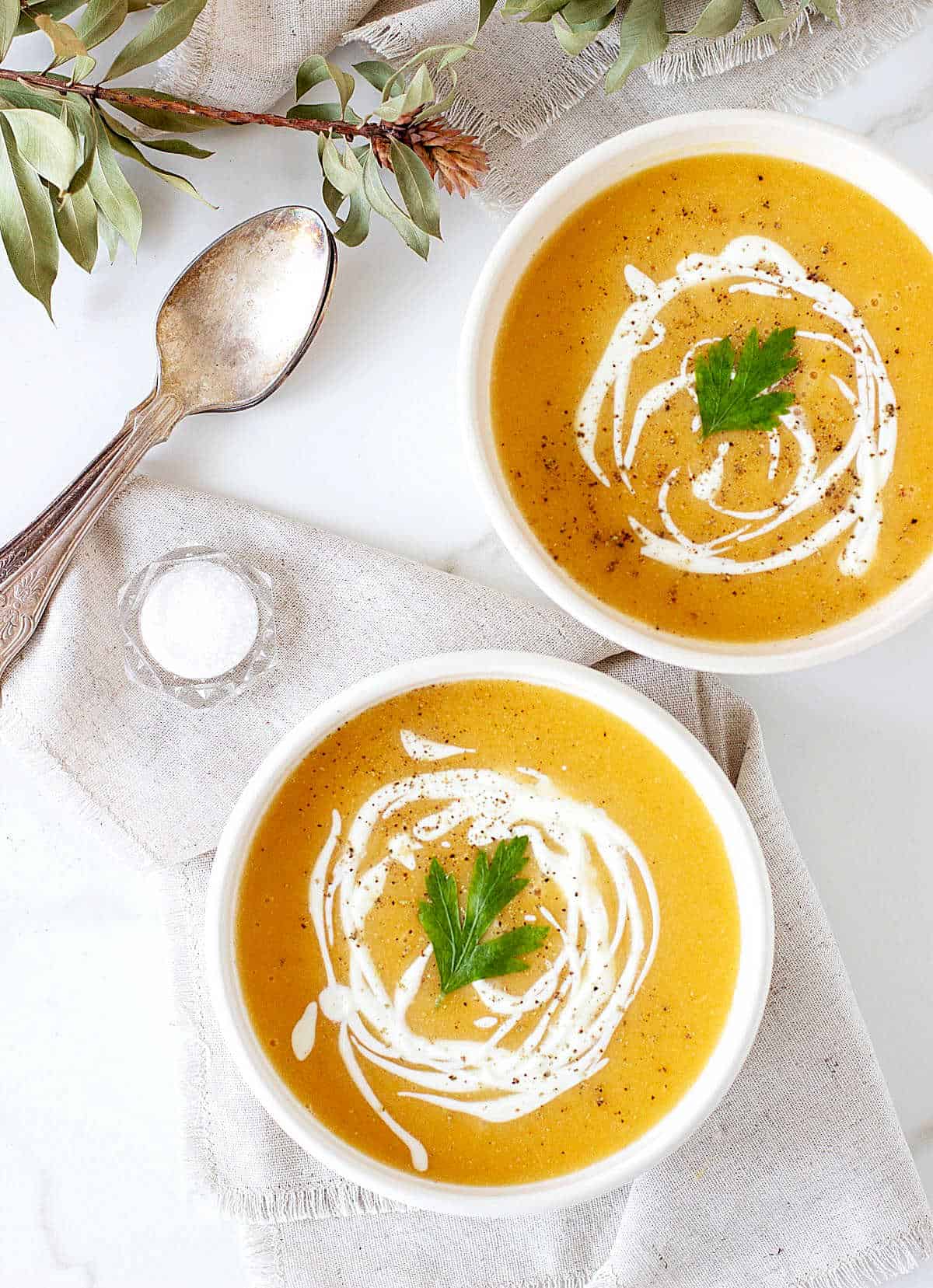 Two white bowls of orange soup, white background, spoons, salt.