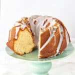 Almond bundt cake with glaze and slice missing on a green cake stand. White background.