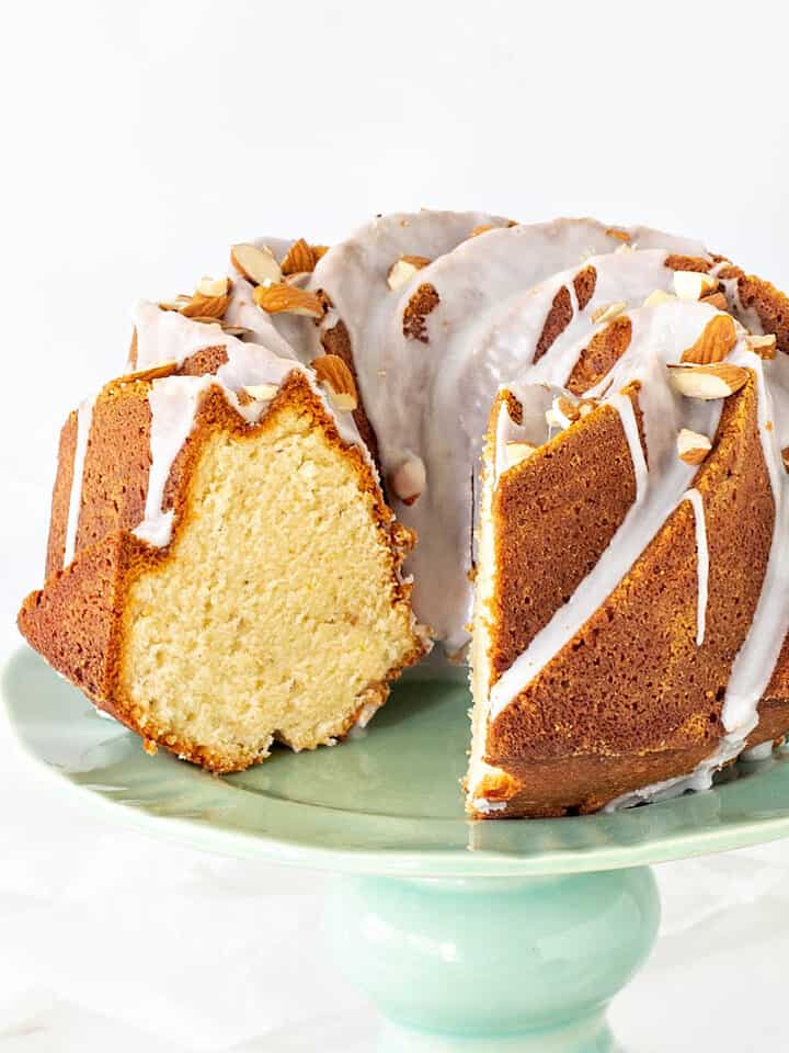 Almond bundt cake with glaze and slice missing on a green cake stand. White background.