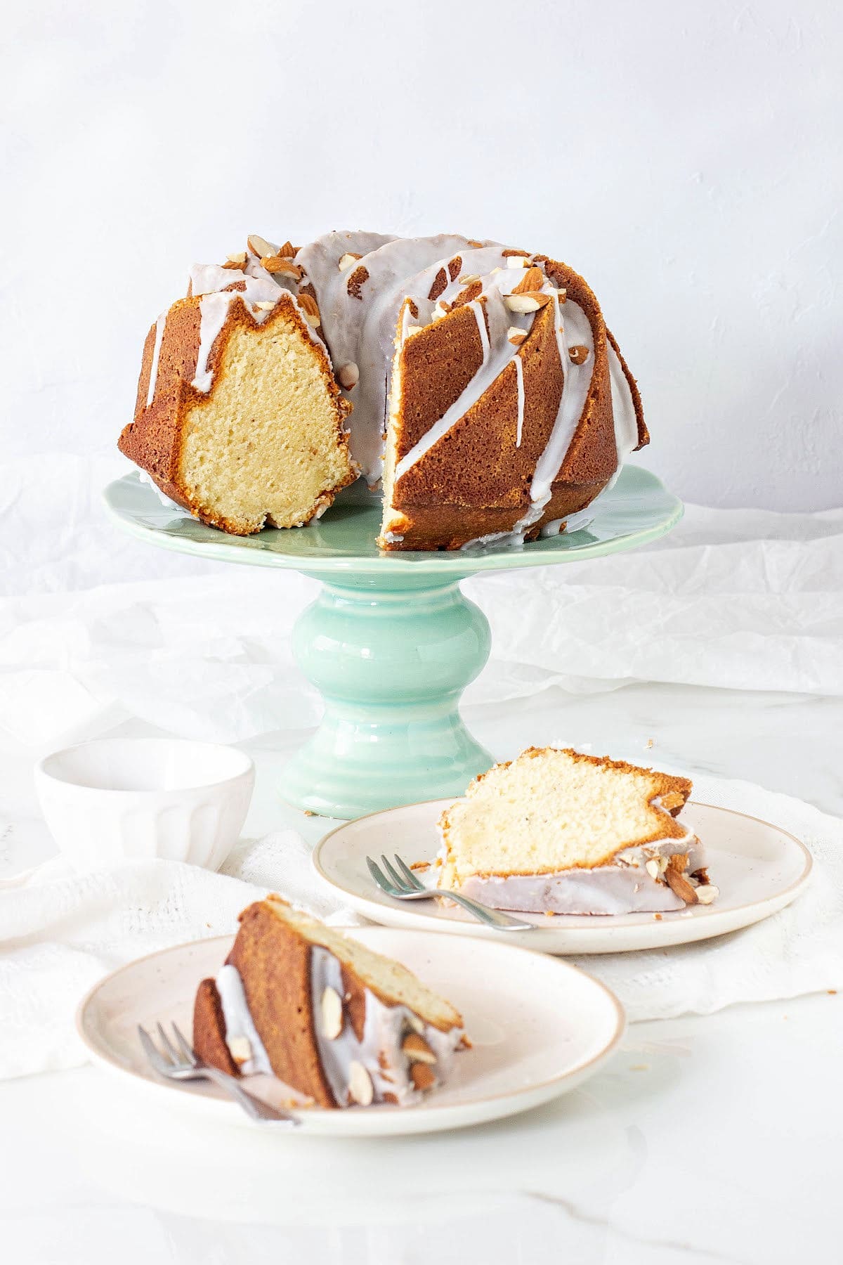 Green cake stand with glazed almond bundt cake. Two slices on plates, white background.