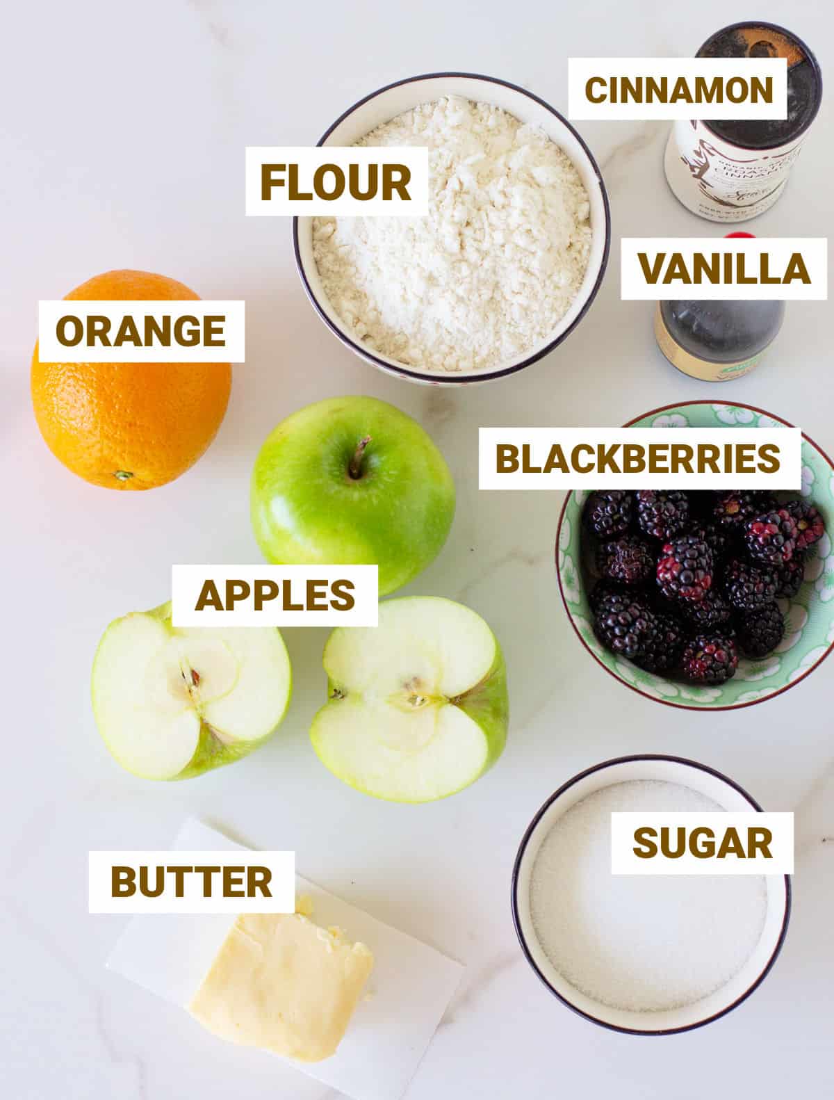 Ingredients for apple blackberry crumble on a white marble surface, including flour, vanilla, orange, butter, sugar.