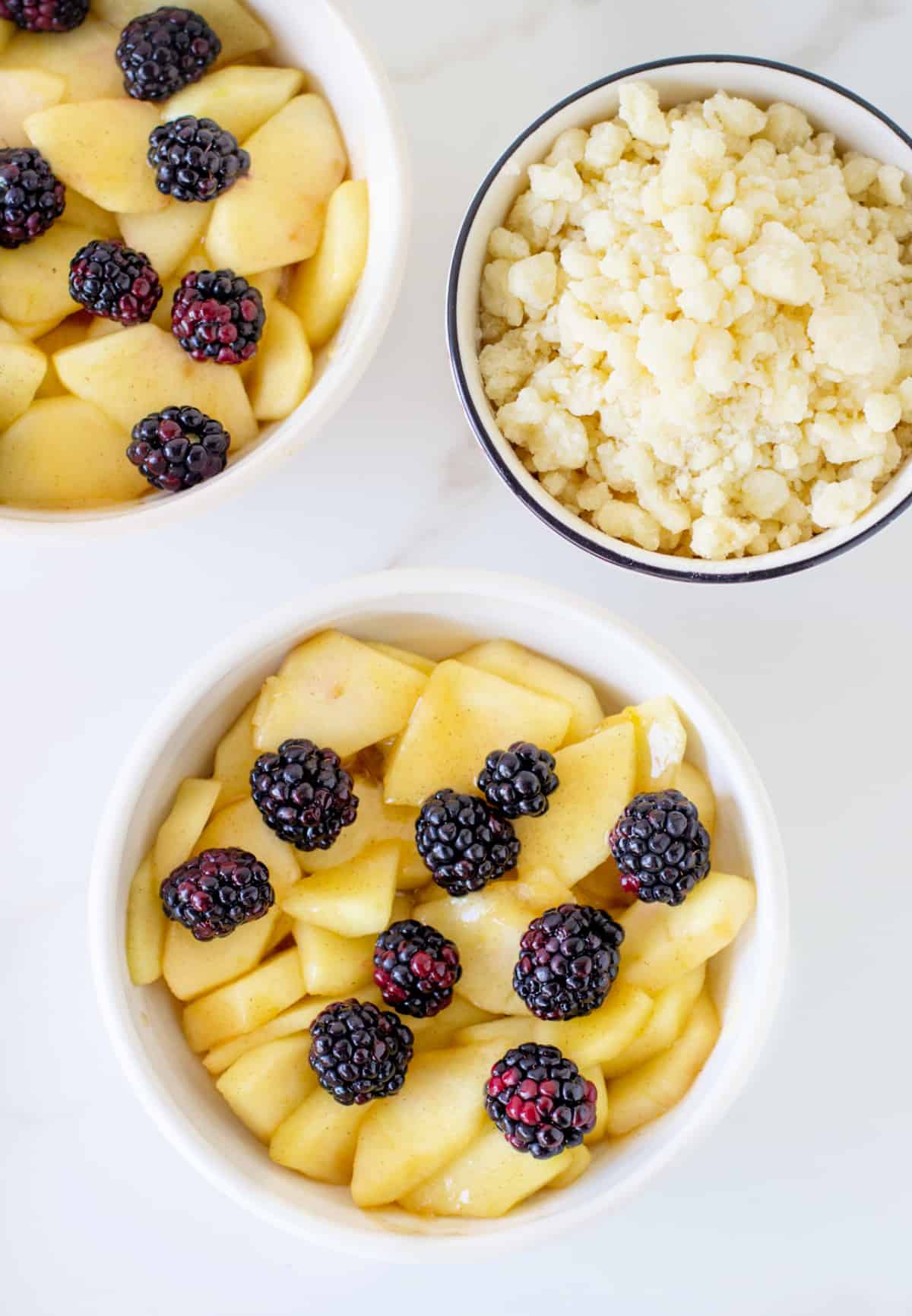 White bowls with apples and blackberries, another with crumble; marble surface.