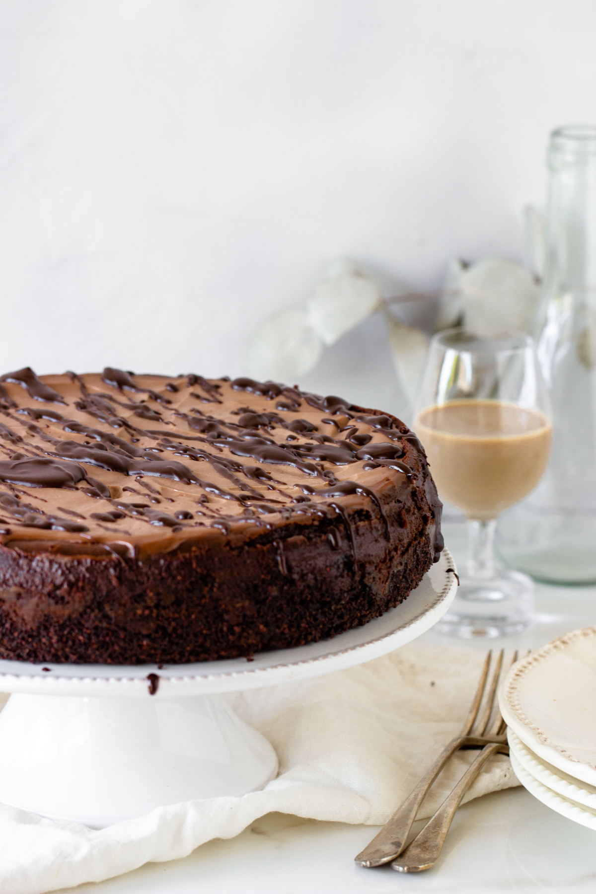 Whole chocolate cheesecake on white cake stand, glass with baileys, forks, piece of cloth