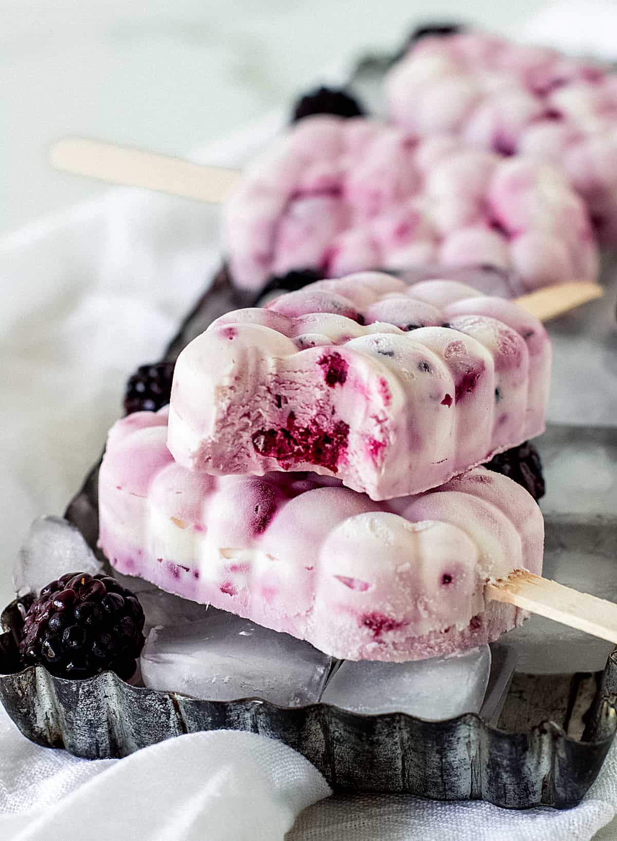 Several berry cheesecake ice cream popsicles, one bitten, on top of a rectangular metal pan