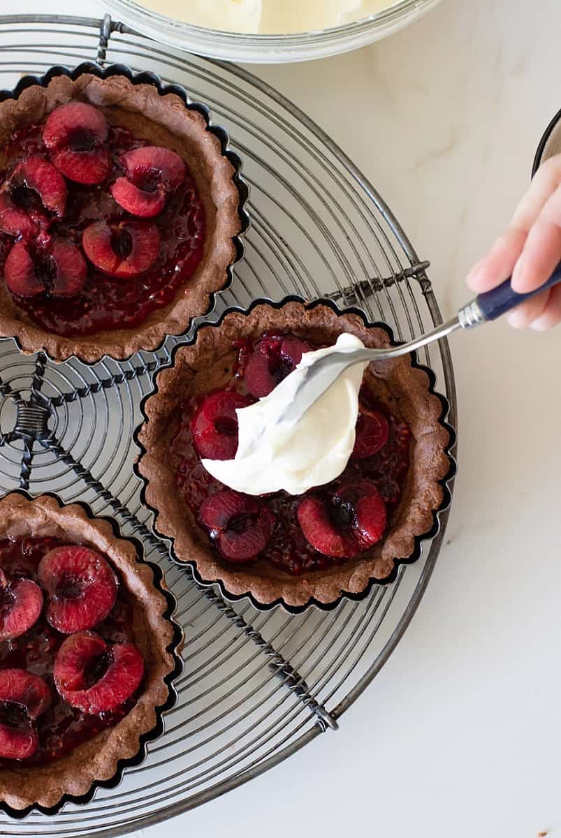 Chocolate cherry tarts on wire rack, hand adding whipped cream with spoon