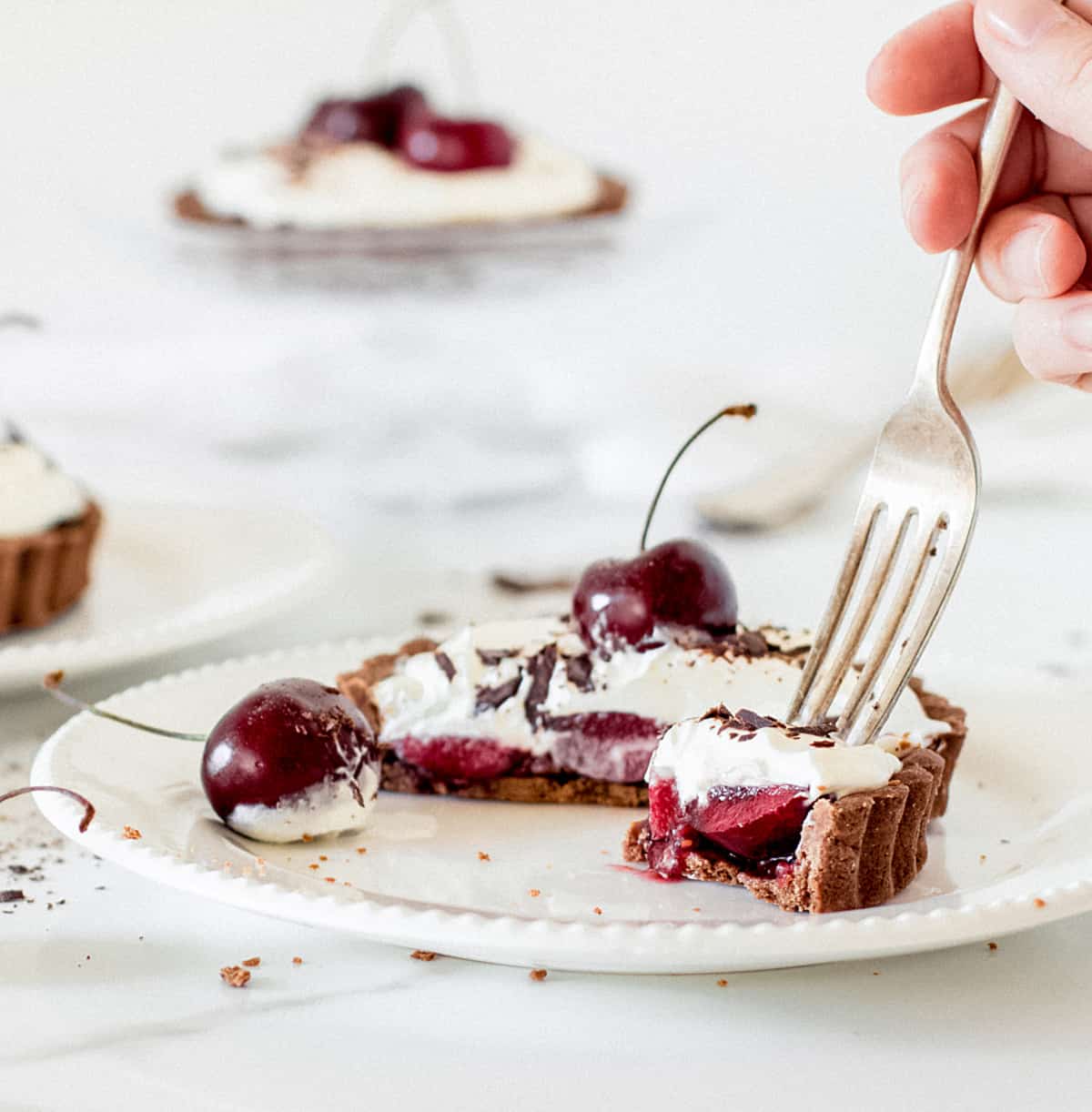 Hand forking piece of chocolate, cherry and cream tartlet, white background, more individual tarts