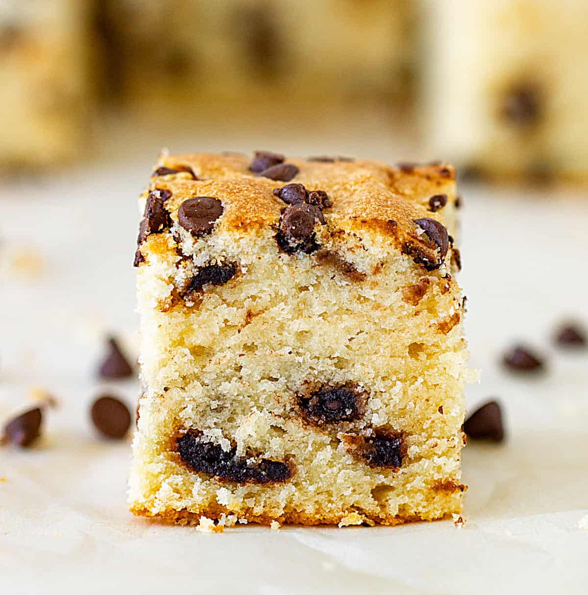 Frontal image of a vanilla cake square studded with chocolate chips on white surface.