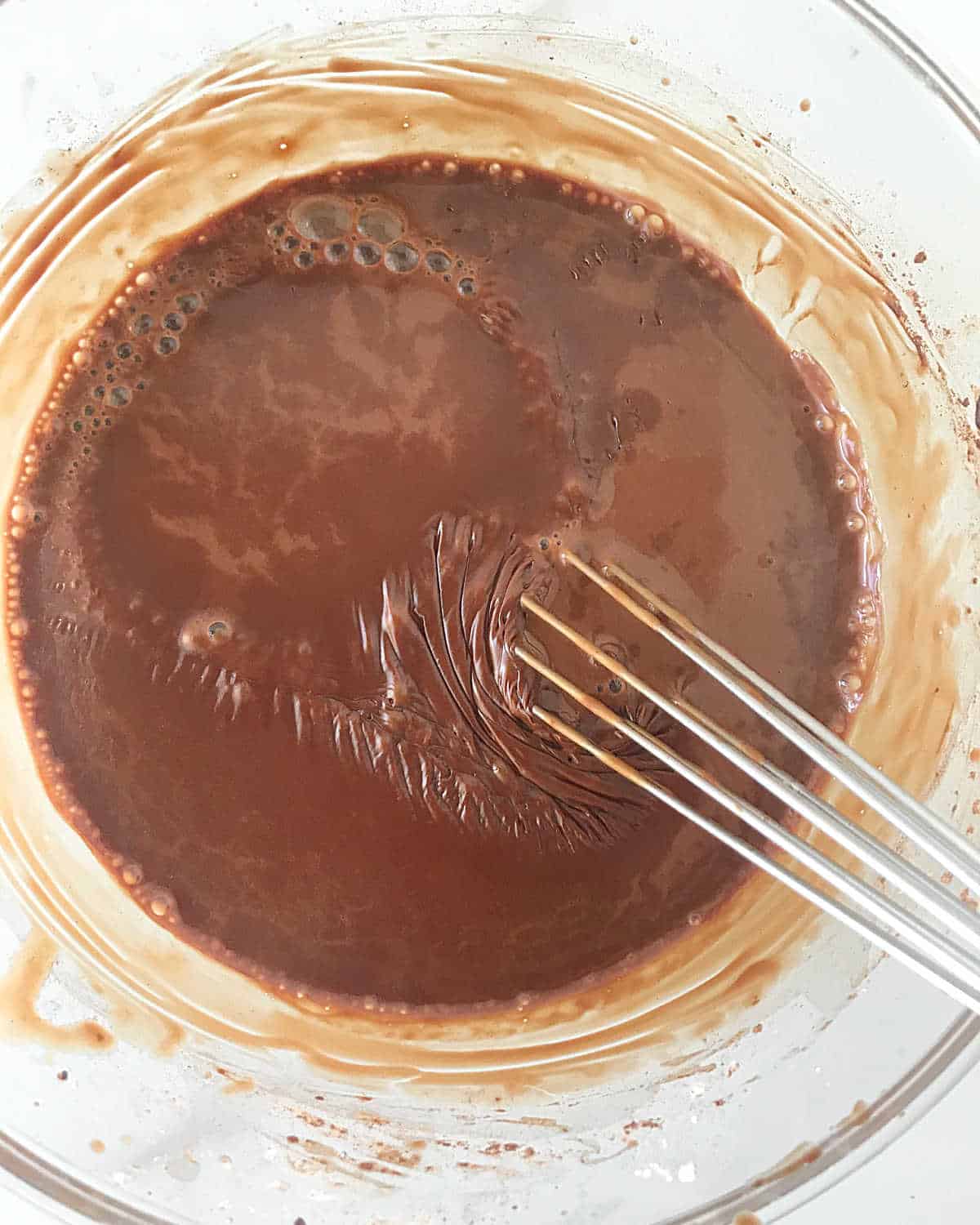 Metal whisk stirring chocolate pudding in a glass bowl.