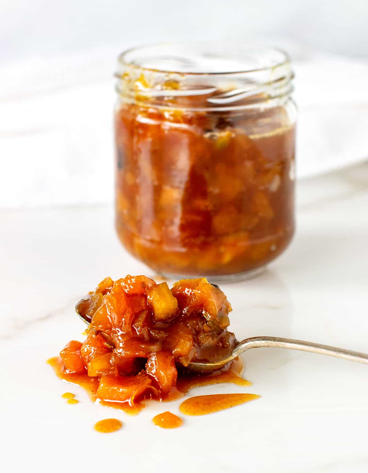 Silver spoon with peach chutney, glass jar in the background, white surface.