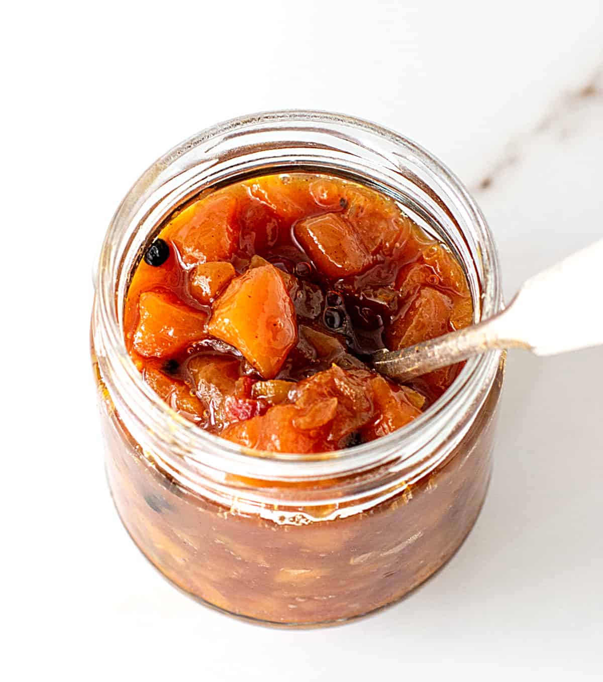 Glass jar with peach chutney, silver spoon inside, white marble surface.