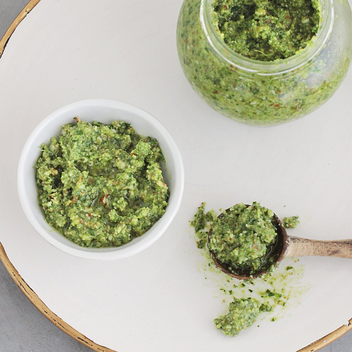 Top view of a gold rimmed white plate with a jar, white bowl and wooden spoon containing pesto