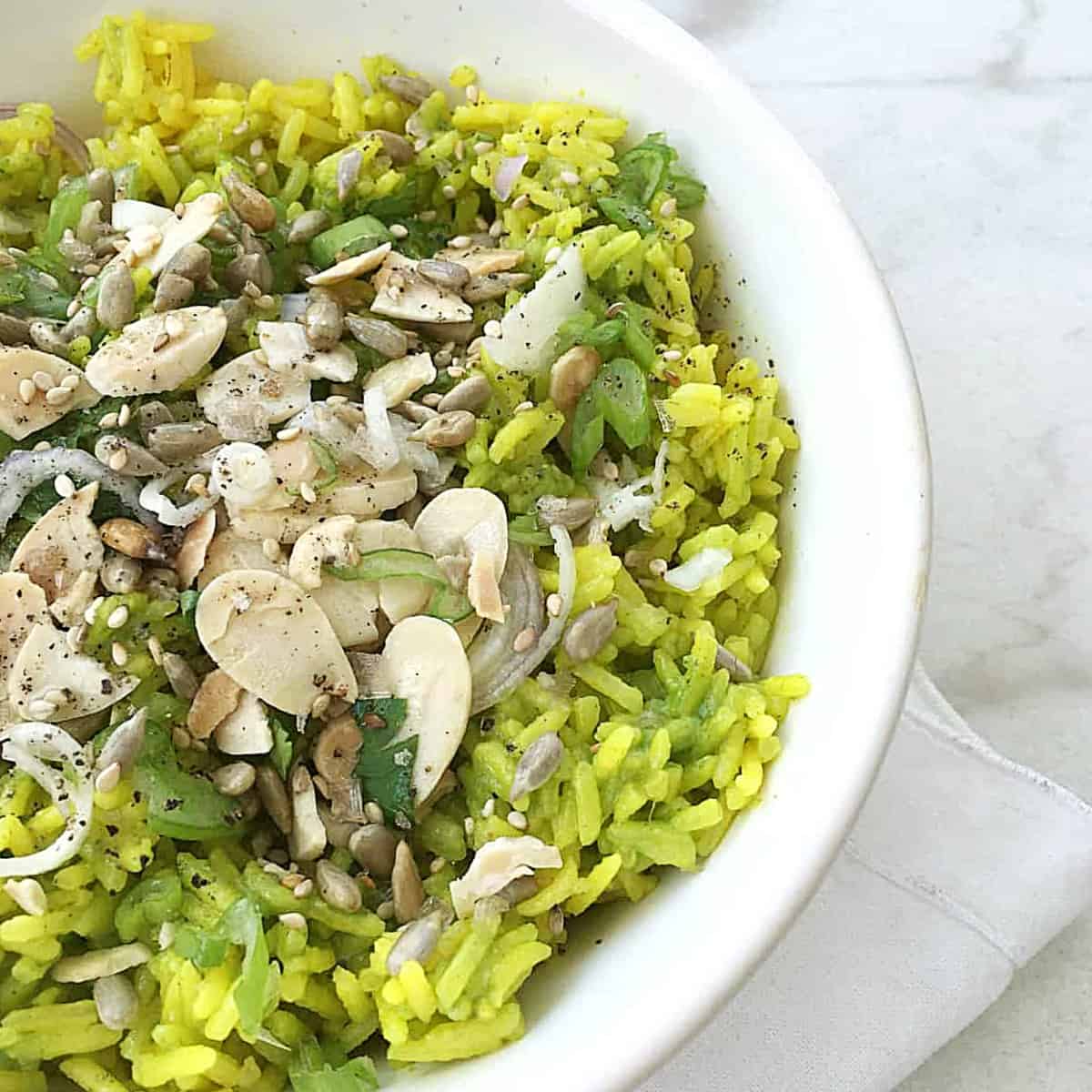 Half image of white bowl with green rice with almonds and seeds on white surface.