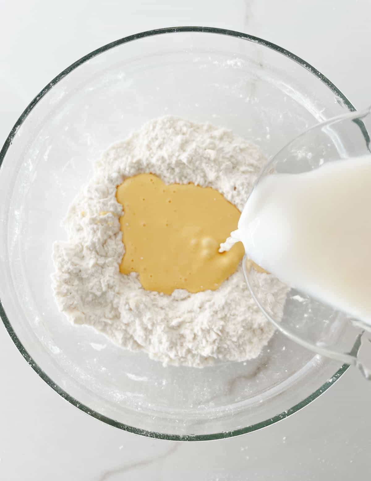 Adding milk and egg to scone dough in a glass bowl on a white surface.