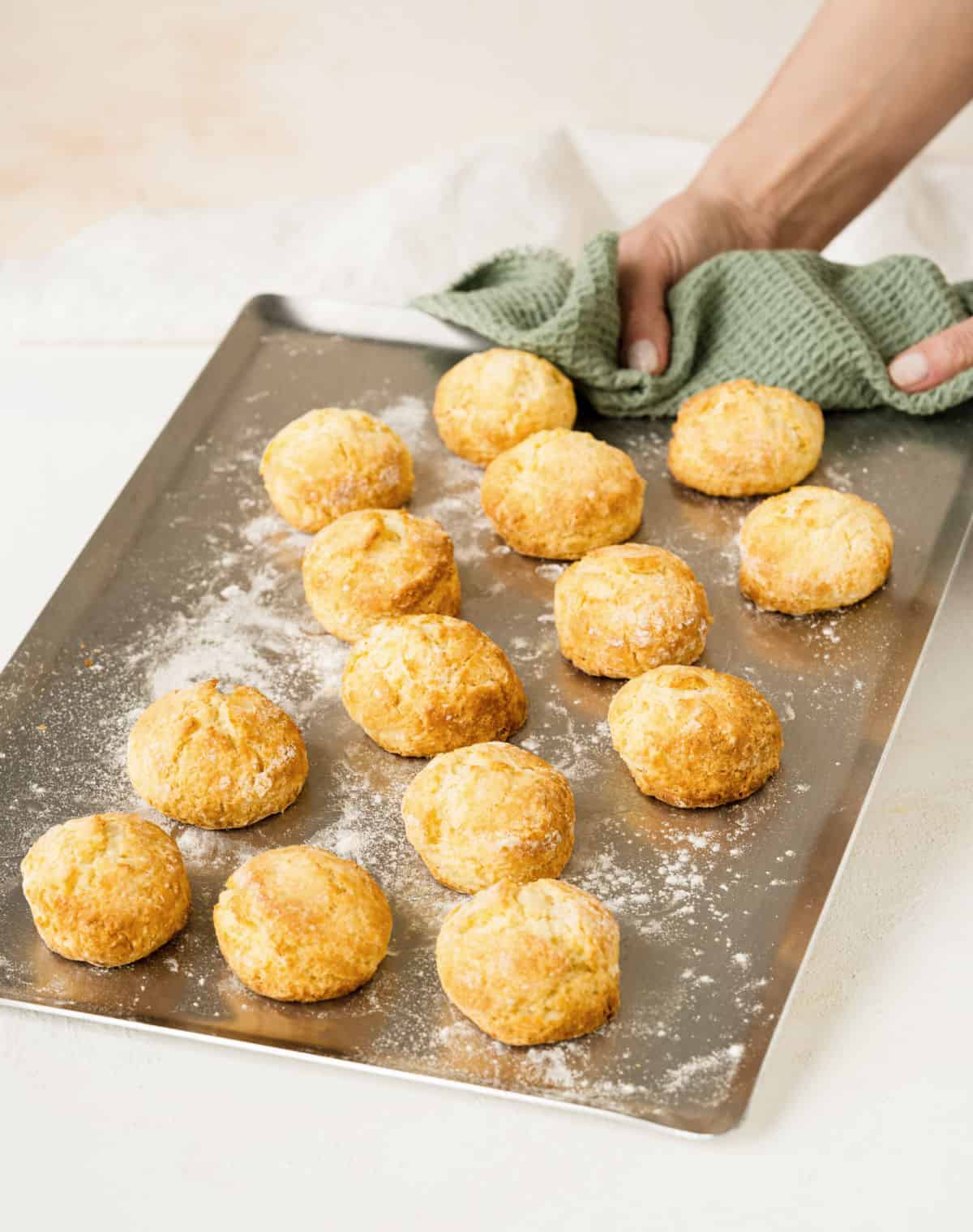 Baked scones on a baking sheet on a white surface. A hand holding it with a green towel.