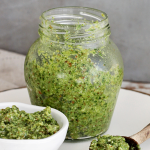 Jar, white bowl and wooden spoon with pesto on gold rimmed white plate, grey background with text