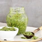 White bowl, jar, and wooden spoon with cilantro pesto on a white plate. Grey background.