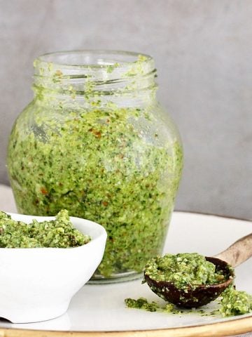 White bowl, jar, and wooden spoon with cilantro pesto on a white plate. Grey background.